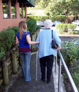 A young woman assists an elderly lady using a walker along a path, surrounded by greenery and parked cars.