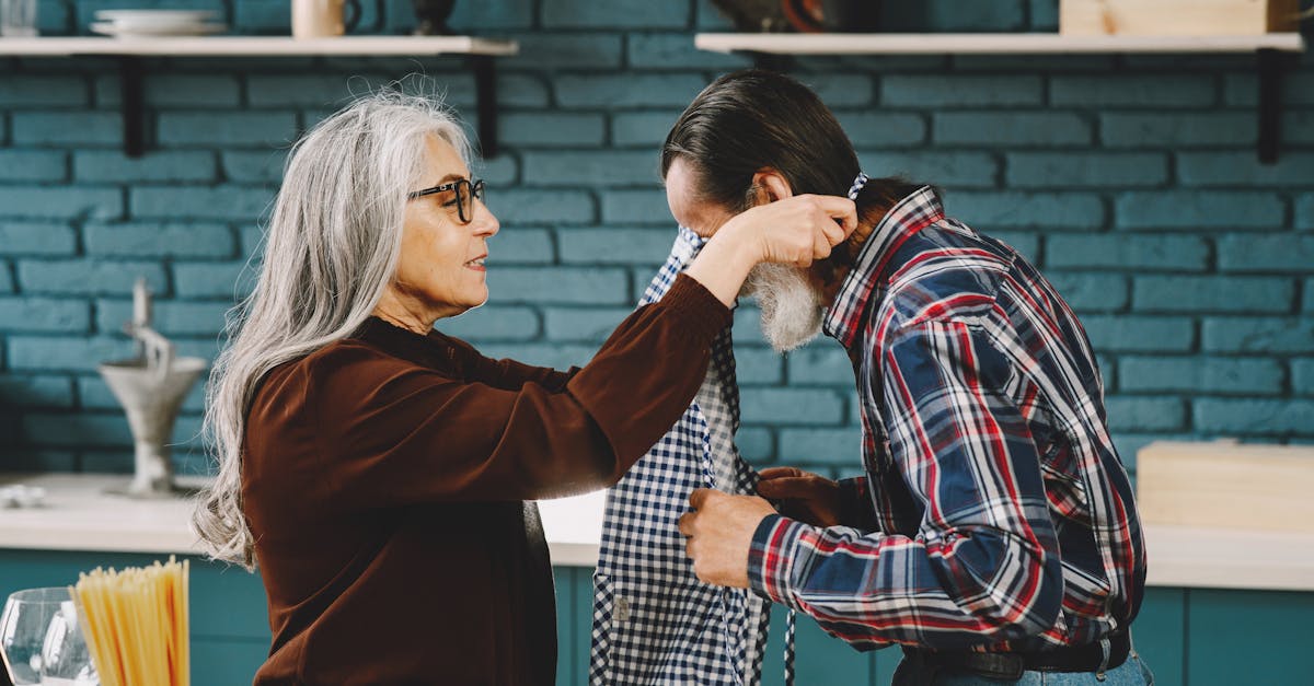 A woman helps a man put on an apron in a cozy kitchen setting with blue brick walls.
